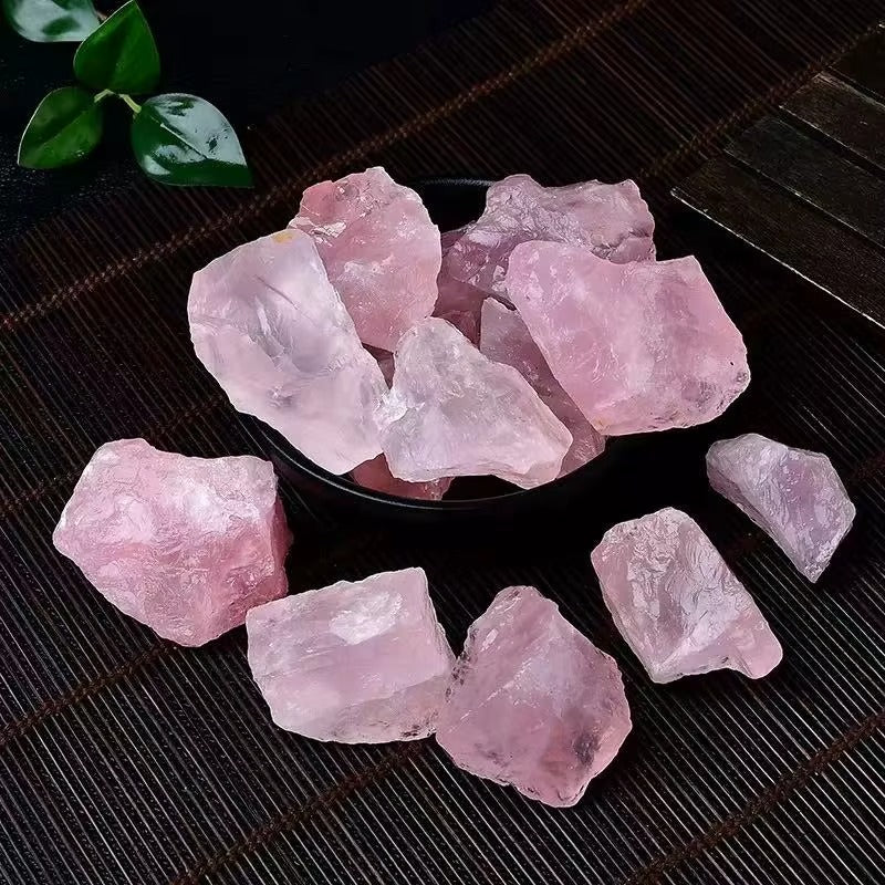 Overhead view of multiple raw rose quartz rough chunks in a dark bowl and scattered on a bamboo mat, soft pink color with waxy luster