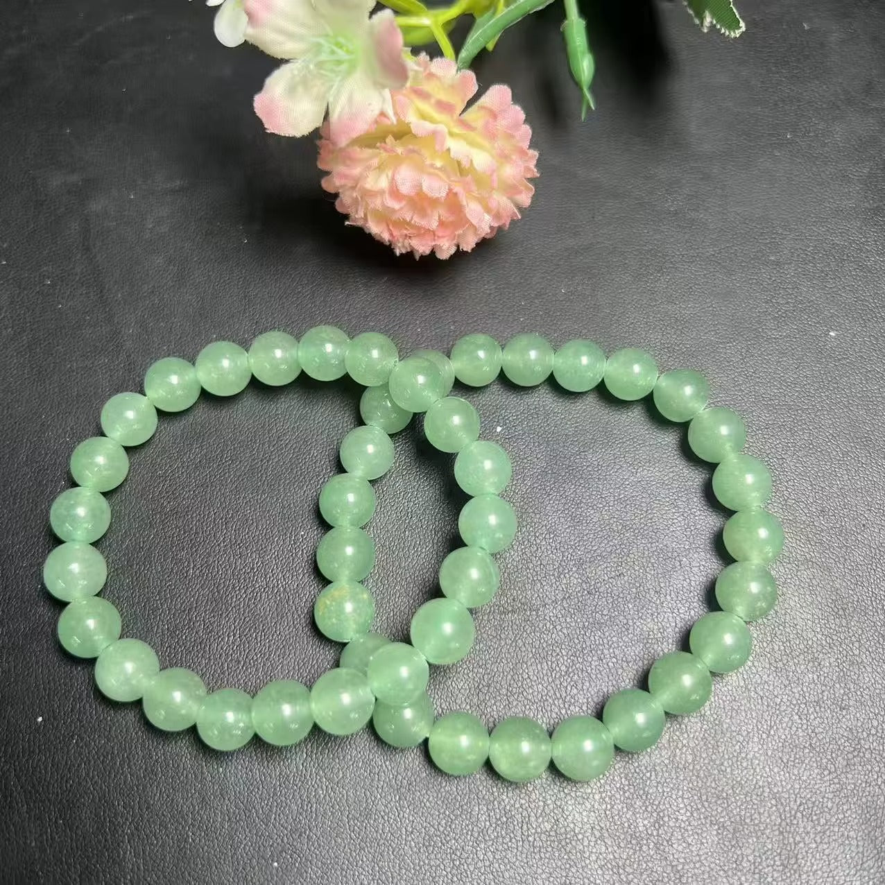 Two green aventurine 8mm bead stretch bracelets overlapping on dark leather surface with pink flower prop, top-down view showing soft apple-green color and vitreous luster