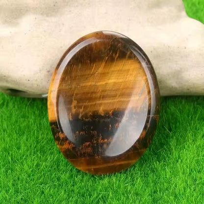 Oval polished Tiger's Eye worry stone showing vivid golden-amber and dark brown chatoyant bands, propped upright on artificial grass with stone backdrop, approximately 2 inches tall