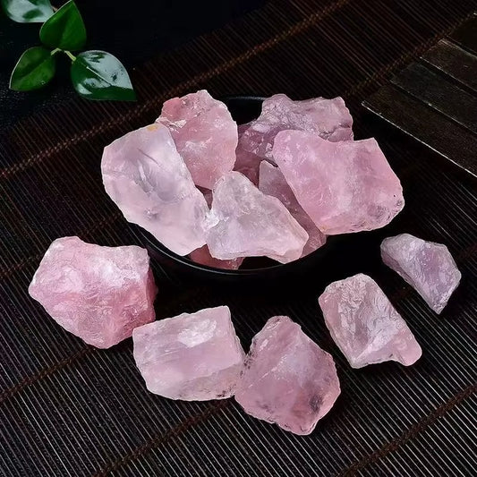 Overhead view of multiple raw rose quartz rough chunks in a dark bowl and scattered on a bamboo mat, soft pink color with waxy luster